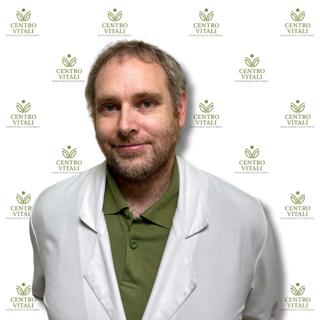 Hombre con barba, delantal blanco y camisa verde, posando frente a un fondo con logotipos de salud mental.